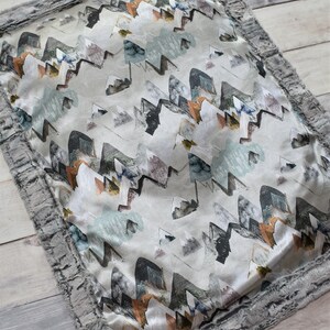 Puede incluir: Una almohada gris y blanca con un patr&oacute;n de formas abstractas en tonos grises, azules, verdes y naranjas. Tiene un ribete de piel sint&eacute;tica gris.