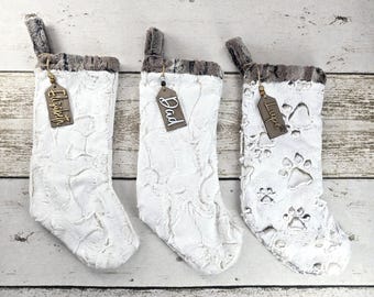 May include: Three white Christmas stockings with faux fur trim and wooden tags. One tag reads "Elizabeth", another "Dad", and the third has paw prints and the name "Luce". The stockings have a textured pattern.