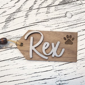 May include: A wooden dog tag with the name "Rex" engraved in silver lettering. A paw print is also engraved on the tag. The tag is attached to a brown twine.