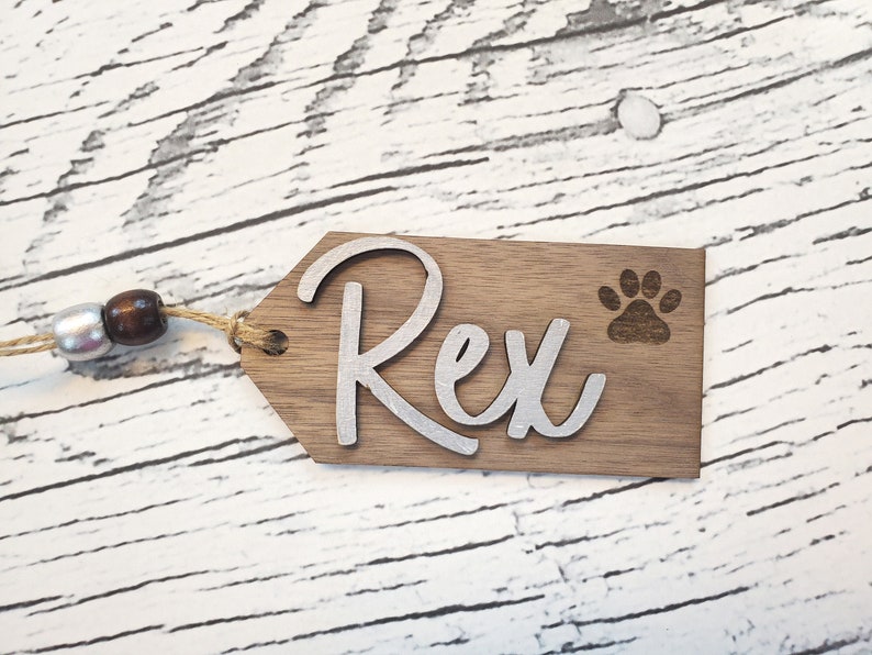 May include: A wooden dog tag with the name "Rex" engraved in silver lettering. A paw print is also engraved on the tag. The tag is attached to a brown twine.