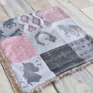 Op de afbeelding: Een roze en grijze patchwork babydeken met een paardenontwerp, een hoefijzer en de tekst "Wild and Free". De deken heeft een zachte, pluche rand.