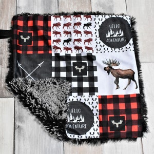 May include: A patchwork baby blanket with a black and white buffalo check pattern, red and black check, and a white background with moose and trees. The blanket has a black and grey faux fur trim. The text "Hello Adventure" is printed on the blanket.