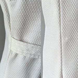 May include: Close-up of a white textured fabric, possibly a jacket or coat. The fabric has a woven pattern, and a small belt detail is visible. The garment appears to be made of a thick, durable material.