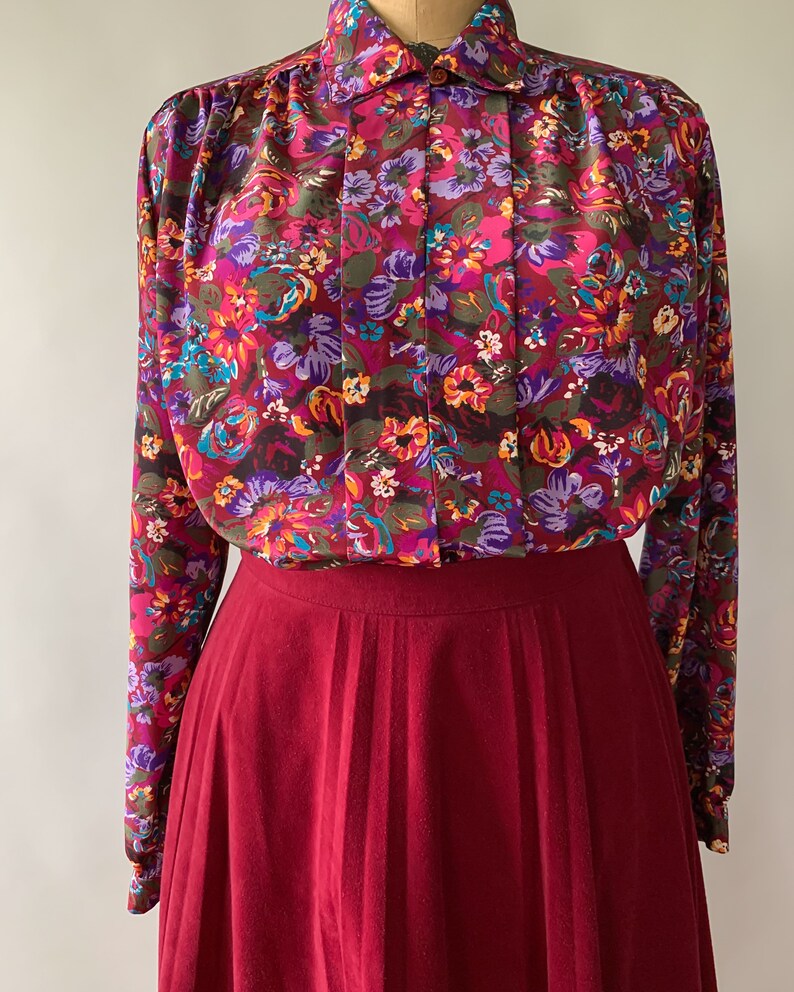 May include: A vintage floral print blouse with a burgundy pleated skirt. The blouse features a vibrant pattern of pink, purple, and orange flowers on a dark background. The skirt is a solid, deep red color.