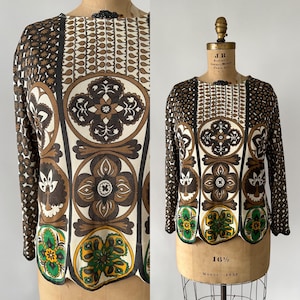 Puede incluir: Top de manga larga con un patrón geométrico único en marrón, crema y verde. El diseño presenta motivos florales y circulares. El top se muestra en un maniquí vintage.