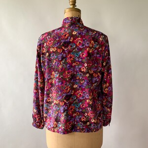 May include: A long-sleeved blouse with a vibrant floral pattern. The blouse features a high collar and is adorned with a mix of purple, pink, orange, and teal flowers on a dark brown background. The garment is displayed on a vintage dress form.