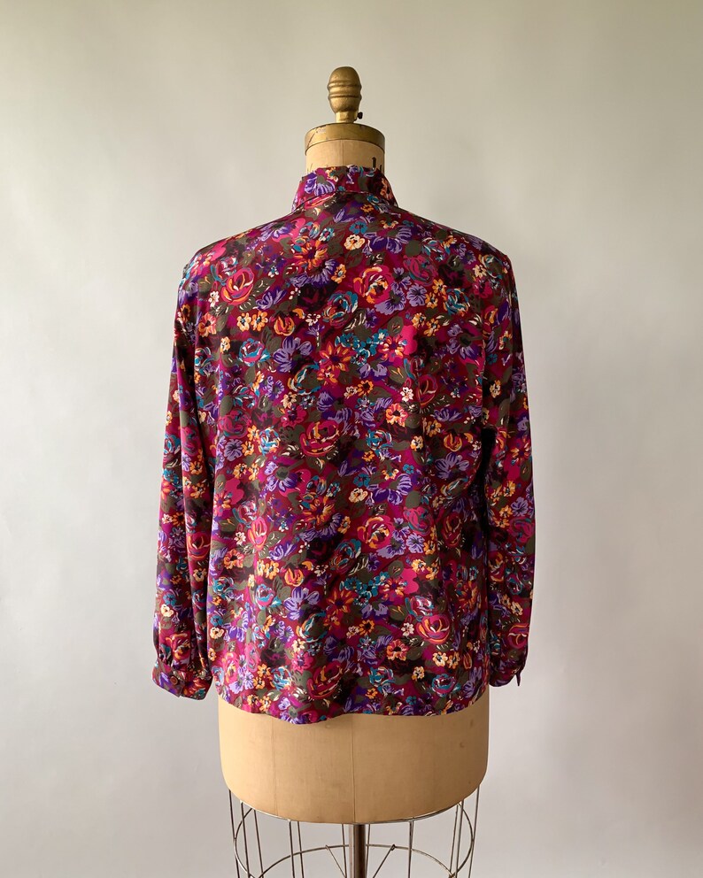 May include: A long-sleeved blouse with a vibrant floral pattern. The blouse features a high collar and is adorned with a mix of purple, pink, orange, and teal flowers on a dark brown background. The garment is displayed on a vintage dress form.