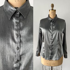 Puede incluir: Una camisa abotonada de manga larga en tejido gris metálico. La camisa tiene un cuello clásico y se muestra en un maniquí vintage. La tela tiene un brillo sutil y los botones son de color gris a juego. La camisa es una prenda versátil.