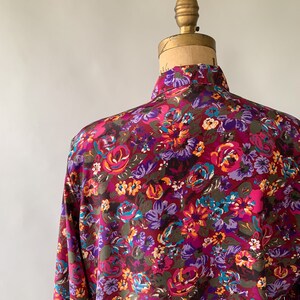 May include: A long-sleeved shirt with a vibrant floral pattern. The shirt features a burgundy background with a colorful array of flowers in shades of pink, purple, orange, and teal. The shirt is displayed on a vintage mannequin.