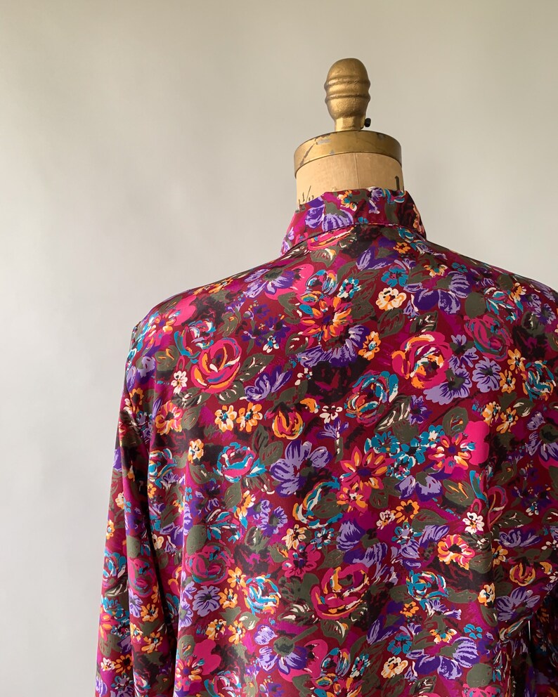 May include: A long-sleeved shirt with a vibrant floral pattern. The shirt features a burgundy background with a colorful array of flowers in shades of pink, purple, orange, and teal. The shirt is displayed on a vintage mannequin.