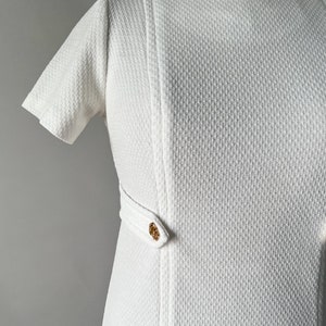 May include: White textured dress with short sleeves and a round neckline. The dress features a vertical seam detail and a decorative gold-toned clasp at the waist. The fabric has a subtle woven pattern, giving it a vintage look.