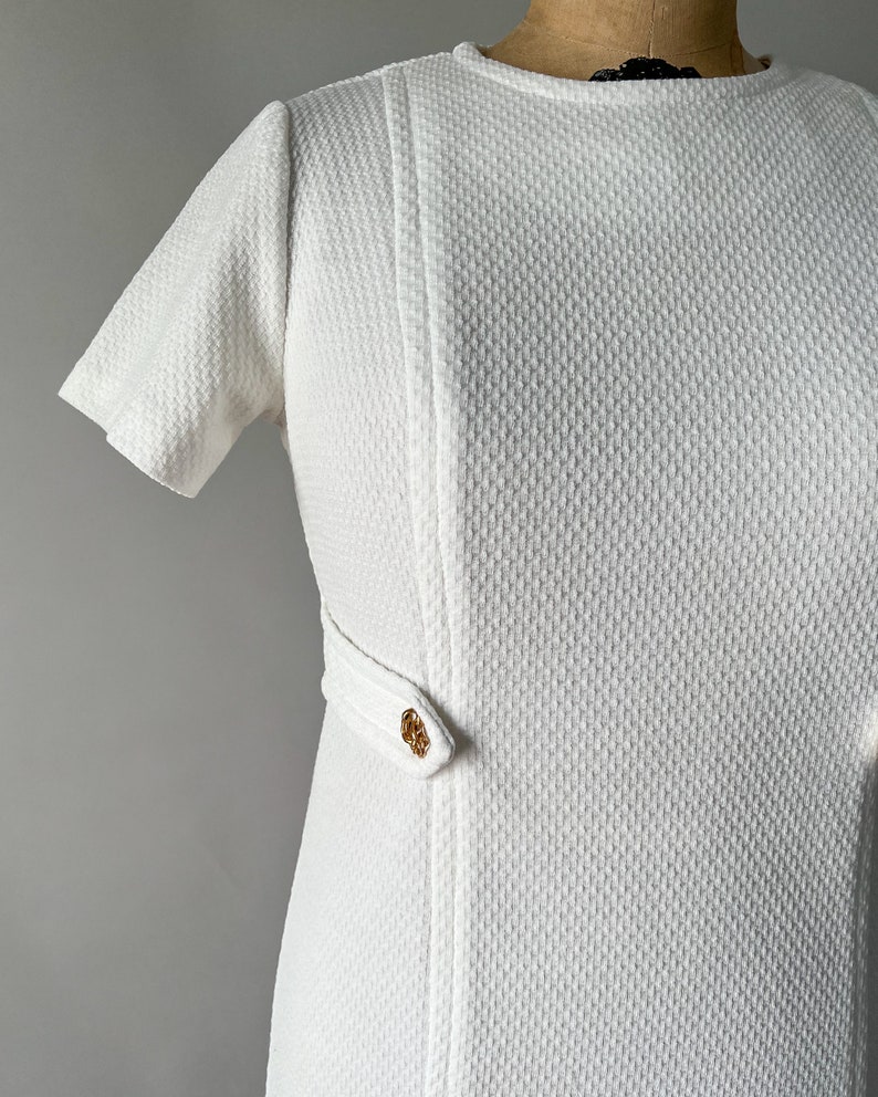 May include: White textured dress with short sleeves and a round neckline. The dress features a vertical seam detail and a decorative gold-toned clasp at the waist. The fabric has a subtle woven pattern, giving it a vintage look.