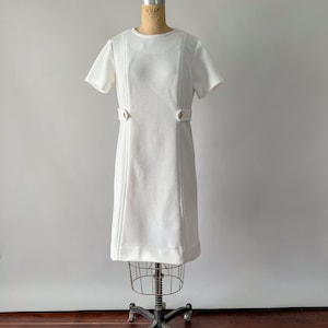 May include: A vintage white dress with short sleeves and a textured fabric. The dress features decorative stitching down the front and small gold-colored buttons at the waist. It is displayed on a vintage dress form.