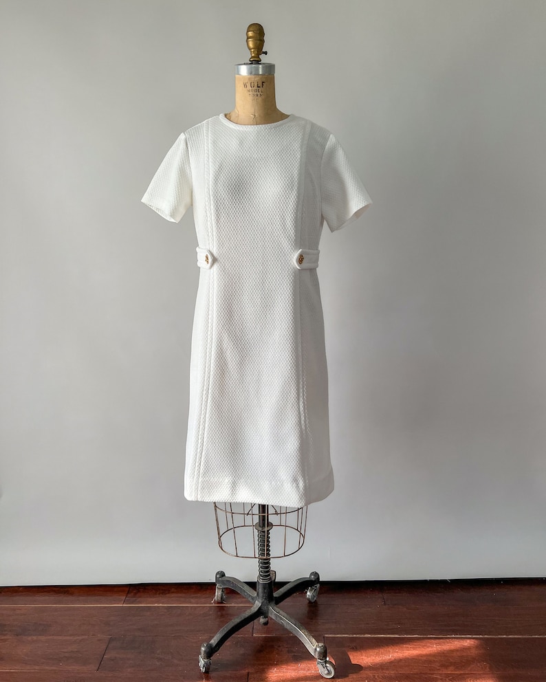 May include: A vintage white dress with short sleeves and a textured fabric. The dress features decorative stitching down the front and small gold-colored buttons at the waist. It is displayed on a vintage dress form.