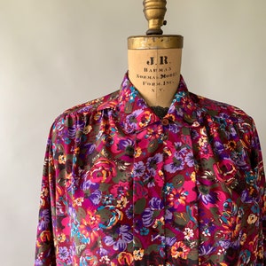 May include: A long-sleeved button-down shirt with a vibrant floral pattern in shades of pink, purple, blue, and yellow. The shirt is displayed on a vintage mannequin. The fabric appears to be a lightweight material.