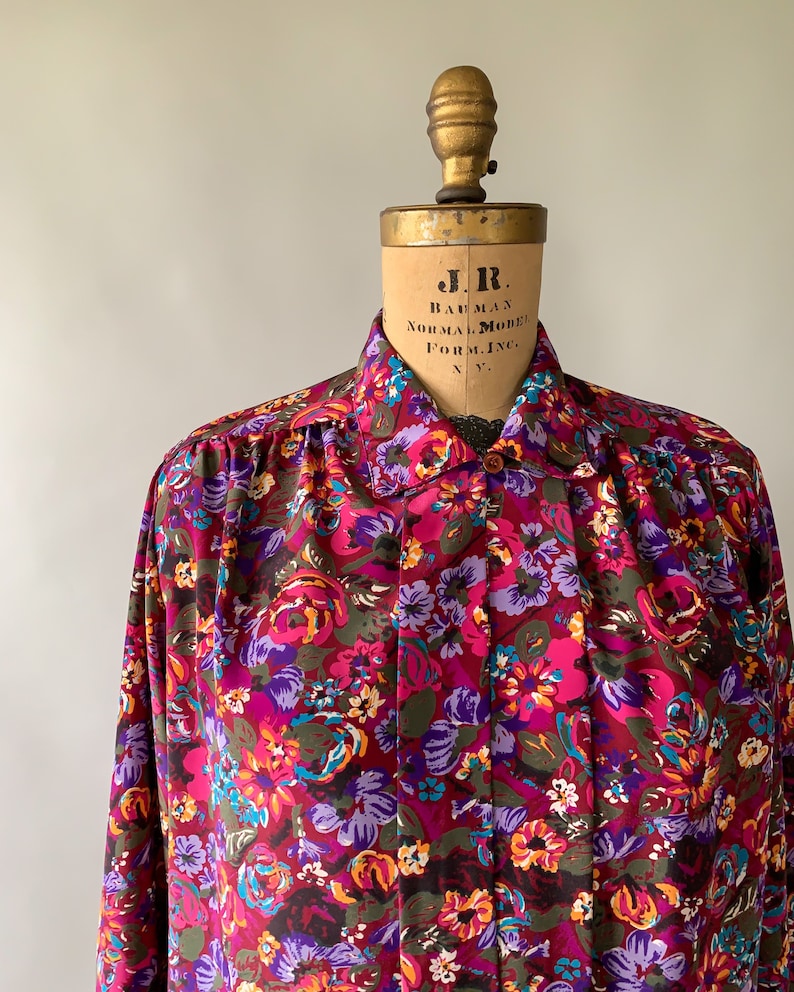 May include: A long-sleeved button-down shirt with a vibrant floral pattern in shades of pink, purple, blue, and yellow. The shirt is displayed on a vintage mannequin. The fabric appears to be a lightweight material.