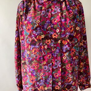 May include: A long-sleeved blouse with a vibrant floral pattern in shades of burgundy, pink, purple, and teal. The blouse features a collared neckline and buttoned cuffs. The fabric is a silky material.