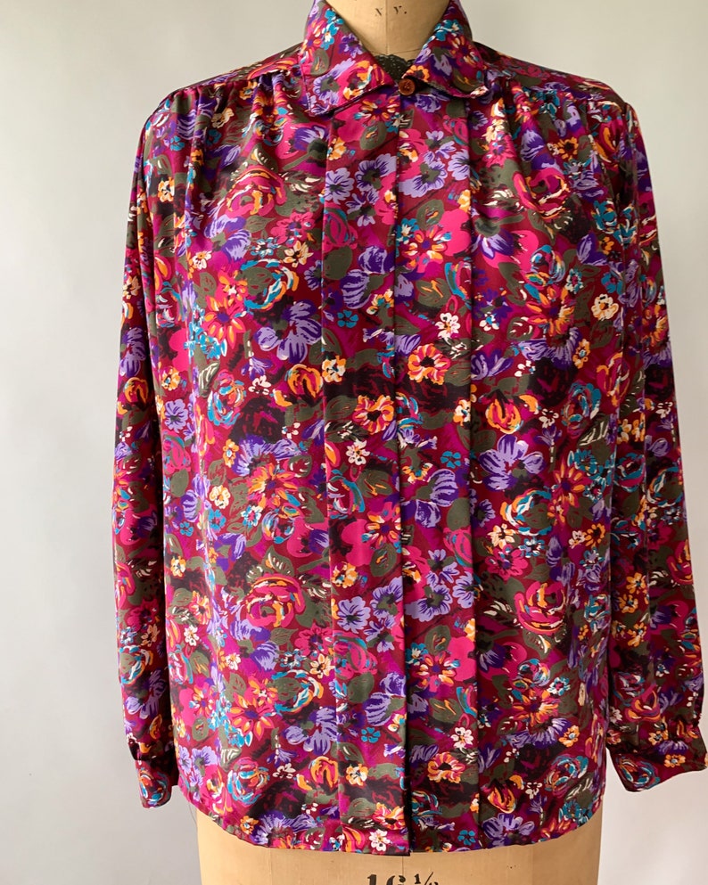 May include: A long-sleeved blouse with a vibrant floral pattern in shades of burgundy, pink, purple, and teal. The blouse features a collared neckline and buttoned cuffs. The fabric is a silky material.