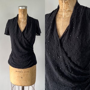 May include: A black wrap top with a V-neckline and short sleeves. The top is made of a sheer fabric with a delicate pattern and is adorned with small, sparkly embellishments.