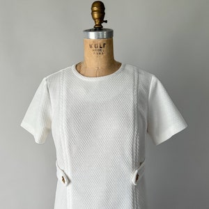 May include: A vintage white dress with short sleeves and a textured fabric. The dress features a round neckline, vertical seams, and decorative gold-colored buttons at the waist. The dress is displayed on a vintage dress form.