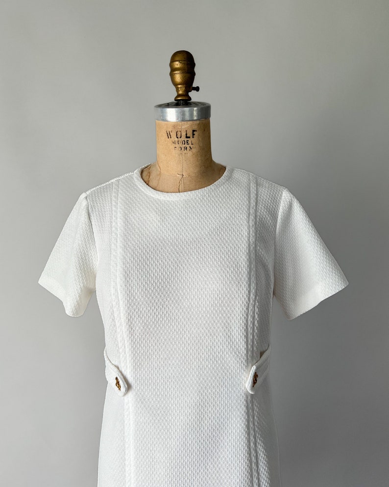 May include: A vintage white dress with short sleeves and a textured fabric. The dress features a round neckline, vertical seams, and decorative gold-colored buttons at the waist. The dress is displayed on a vintage dress form.