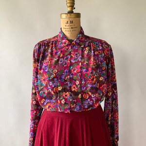 May include: A vintage floral print blouse with a burgundy skirt. The blouse features a vibrant pattern of pink, purple, and orange flowers on a dark background. The skirt is a solid, pleated burgundy color, creating a classic, feminine look.
