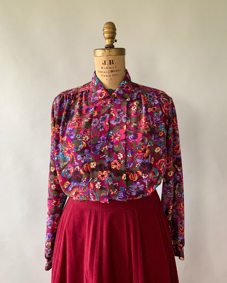 May include: A vintage floral print blouse with a burgundy skirt. The blouse features a vibrant pattern of pink, purple, and orange flowers on a dark background. The skirt is a solid, pleated burgundy color, creating a classic, feminine look.
