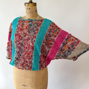 May include: A colorful, textured knit top with a boat neck and wide sleeves. The top features a mix of red, pink, orange, and blue yarns, with turquoise and pink stripes. The garment is displayed on a vintage dress form.