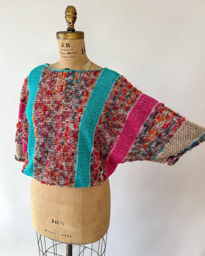 May include: A colorful, textured knit top with a boat neck and wide sleeves. The top features a mix of red, pink, orange, and blue yarns, with turquoise and pink stripes. The garment is displayed on a vintage dress form.