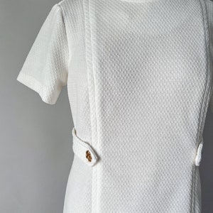May include: A vintage white dress with short sleeves and a textured fabric. The dress features a round neckline and decorative gold-colored buttons on the waist. The dress is displayed on a mannequin.
