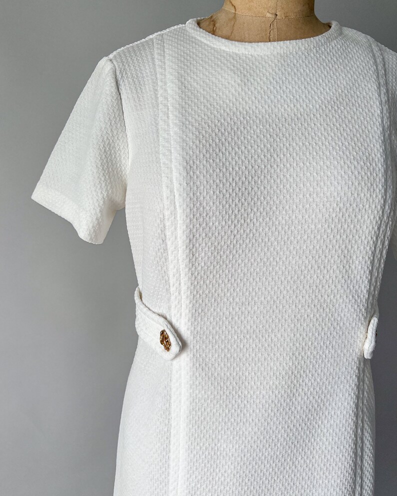 May include: A vintage white dress with short sleeves and a textured fabric. The dress features a round neckline and decorative gold-colored buttons on the waist. The dress is displayed on a mannequin.
