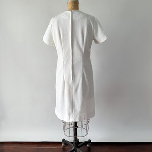 May include: A vintage white dress with short sleeves and a textured fabric. The dress is displayed on a dress form, showcasing its tailored fit and design. The dress has a simple, elegant silhouette, suitable for various occasions.
