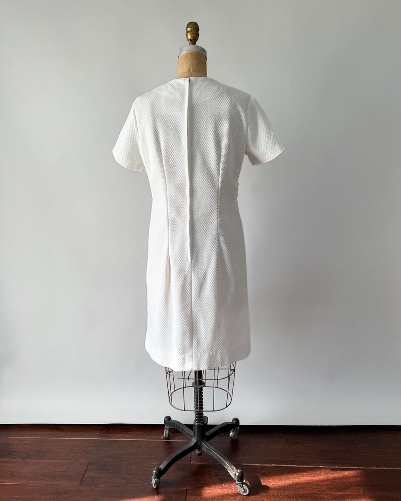 May include: A vintage white dress with short sleeves and a textured fabric. The dress is displayed on a dress form, showcasing its tailored fit and design. The dress has a simple, elegant silhouette, suitable for various occasions.