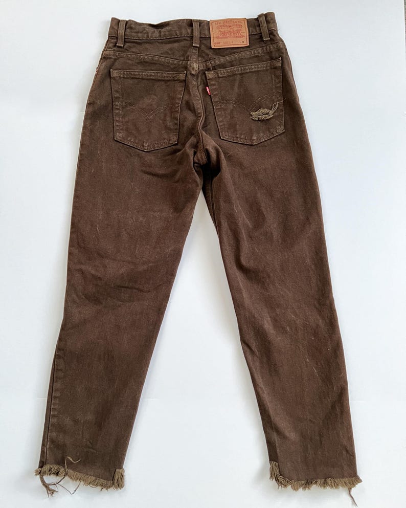 Puede incluir: Vaqueros de mezclilla marr&oacute;n con dobladillos deshilachados. Los vaqueros tienen un peque&ntilde;o parche bordado en el bolsillo trasero que dice "Levi's" y "Original  Quality Clothing".