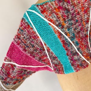 May include: A colorful, textured knit top with a unique design. The garment features a mix of pink, teal, and white accents. The sleeves are a mix of textures and colors, with a white trim. The top is a vibrant piece of wearable art.