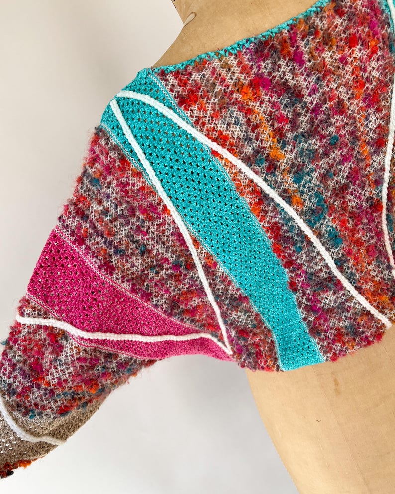May include: A colorful, textured knit top with a unique design. The garment features a mix of pink, teal, and white accents. The sleeves are a mix of textures and colors, with a white trim. The top is a vibrant piece of wearable art.