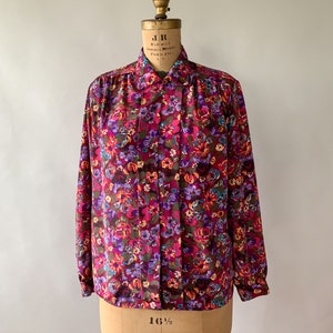 May include: A long-sleeved blouse with a vibrant floral pattern in shades of burgundy, purple, and orange. The blouse is displayed on a vintage dress form, showcasing its button-down front and collared neckline. The fabric appears to be a lightweight material.