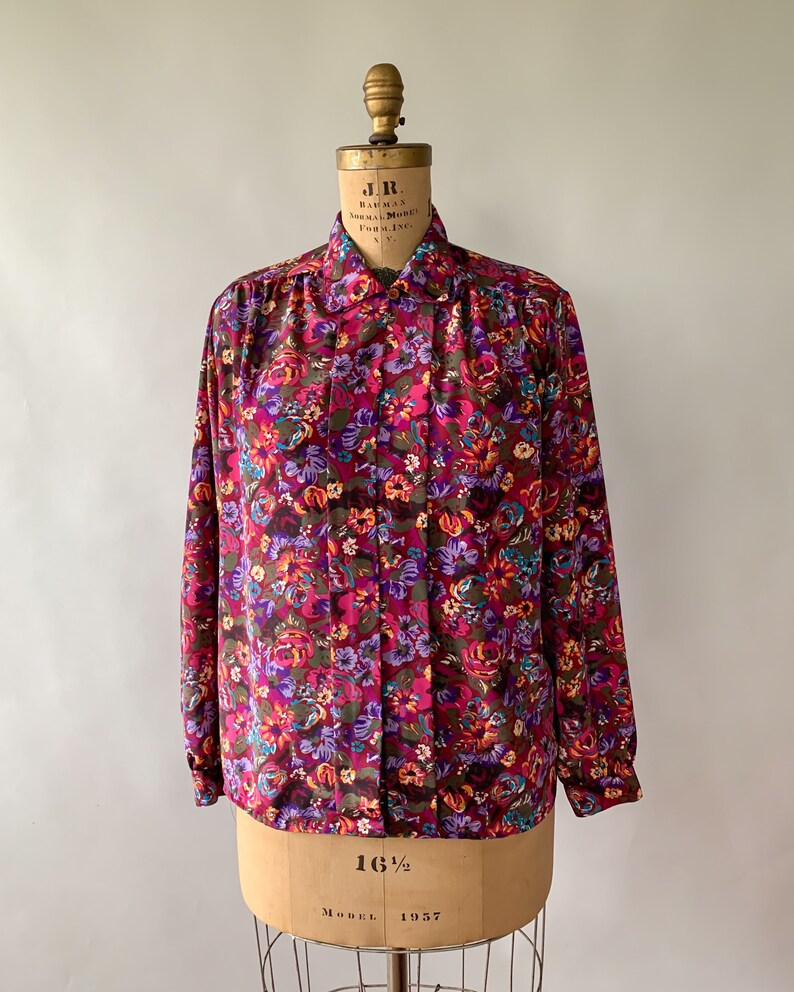 May include: A long-sleeved blouse with a vibrant floral pattern in shades of burgundy, purple, and orange. The blouse is displayed on a vintage dress form, showcasing its button-down front and collared neckline. The fabric appears to be a lightweight material.