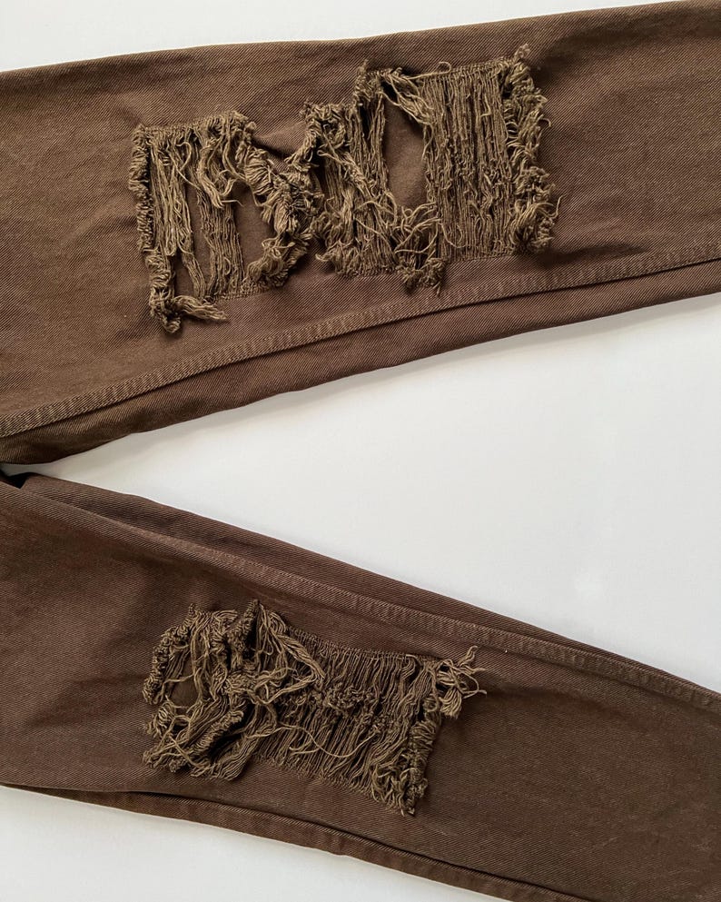 Puede incluir: Un par de pantalones vaqueros de color marr&oacute;n desgastados con bordes deshilachados en las rodillas.