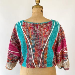 May include: A colorful, textured sweater with a round neckline and short sleeves. The sweater features a mix of vibrant colors, including pink, teal, orange, and brown. White and teal stripes accent the front, and the sleeves have a beige trim. The garment is displayed on a mannequin.