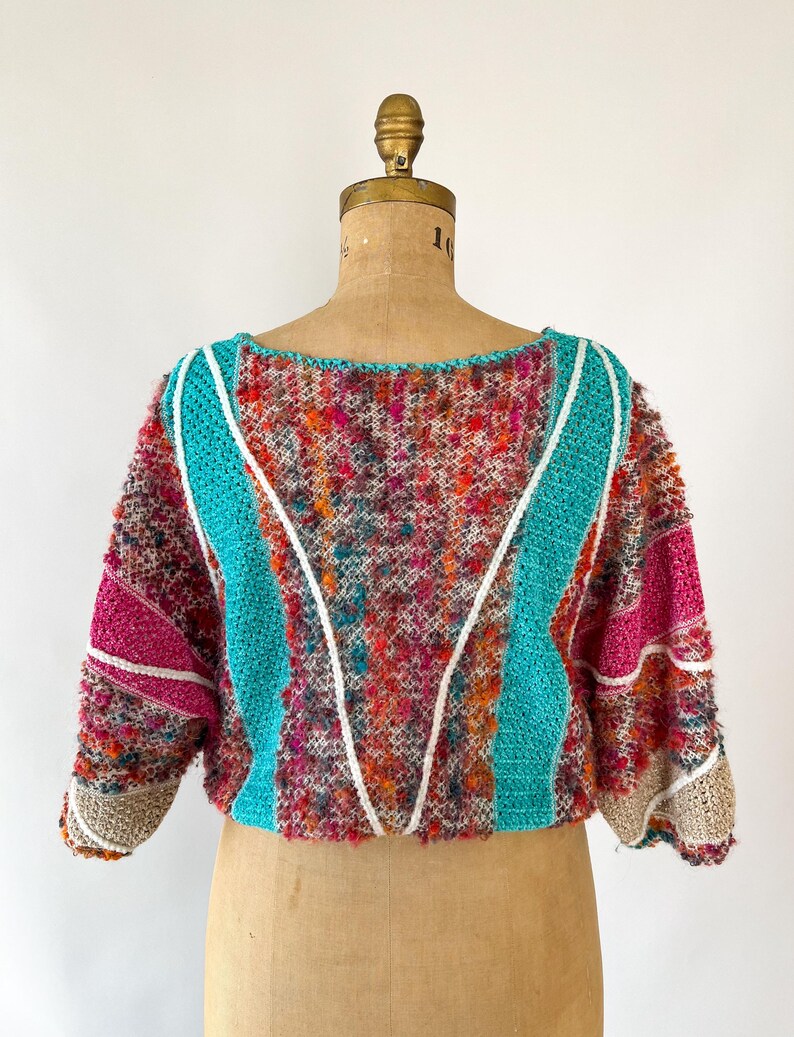 May include: A colorful, textured sweater with a round neckline and short sleeves. The sweater features a mix of vibrant colors, including pink, teal, orange, and brown. White and teal stripes accent the front, and the sleeves have a beige trim. The garment is displayed on a mannequin.
