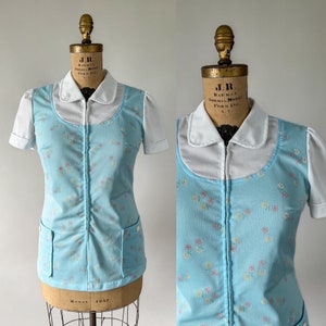 May include: A light blue zip-up top with a white collar and short sleeves. The top has a floral pattern and two pockets. The top is on a mannequin.