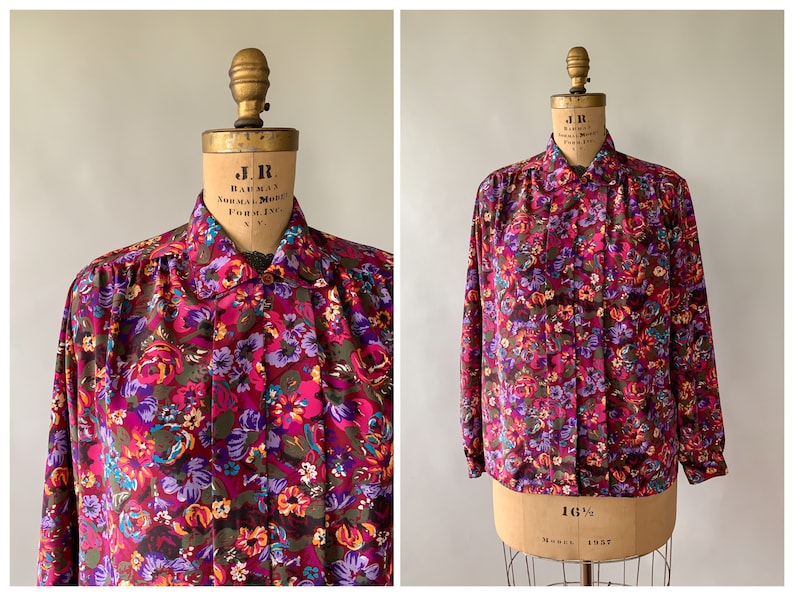 May include: A long-sleeved button-down shirt with a vibrant floral pattern in shades of pink, purple, and green. The shirt is displayed on a vintage mannequin, showcasing its classic design and fit. The fabric appears to be lightweight.