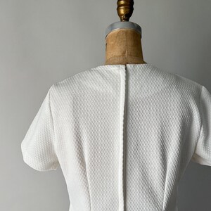 May include: A white, textured dress with short sleeves, displayed on a vintage dress form. The dress has a subtle pattern and a simple, elegant design, perfect for a classic look.