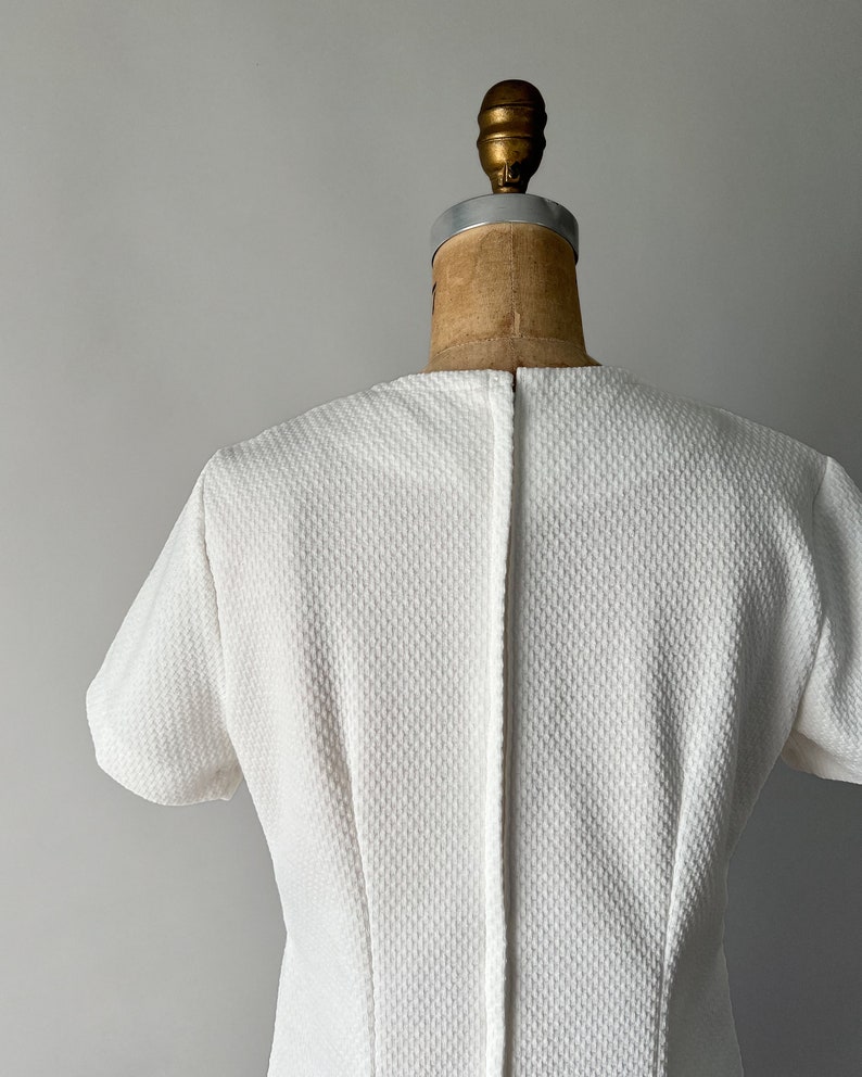 May include: A white, textured dress with short sleeves, displayed on a vintage dress form. The dress has a subtle pattern and a simple, elegant design, perfect for a classic look.