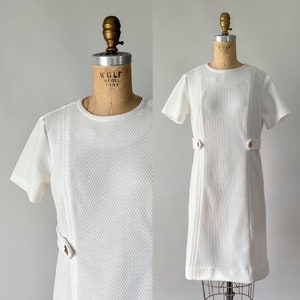 May include: A vintage white dress with short sleeves and a textured fabric. The dress features decorative seams and gold-colored button details at the waist. The dress is displayed on a vintage dress form.