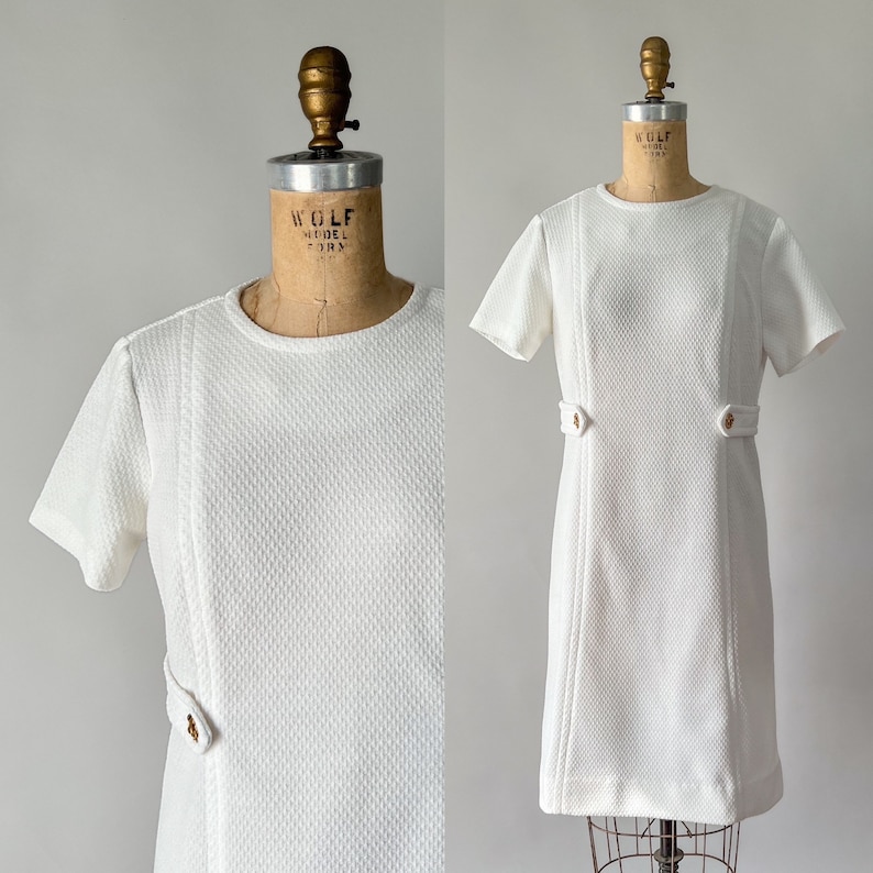 May include: A vintage white dress with short sleeves and a textured fabric. The dress features decorative seams and gold-colored button details at the waist. The dress is displayed on a vintage dress form.