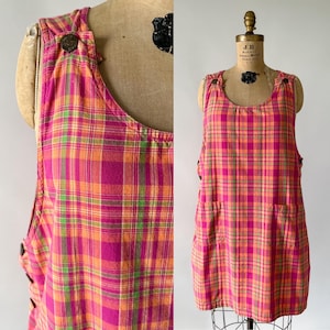 May include: A sleeveless plaid dress in shades of pink, orange, and green, displayed on a mannequin. The dress features a scoop neckline, two front pockets, and decorative buttons on the straps. The fabric has a classic plaid pattern.