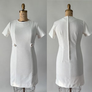 May include: A vintage white dress with short sleeves and a textured fabric. The dress features decorative seams and two gold-colored buttons at the waist. The dress is displayed on a vintage dress form.