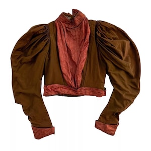 Puede incluir: Una chaqueta marrón recortada con mangas largas y forro de satén rojo. La chaqueta tiene un cuello alto y una cintura ajustada.